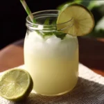 A glass of Brazilian Lemonade served with fresh limes and mint leaves